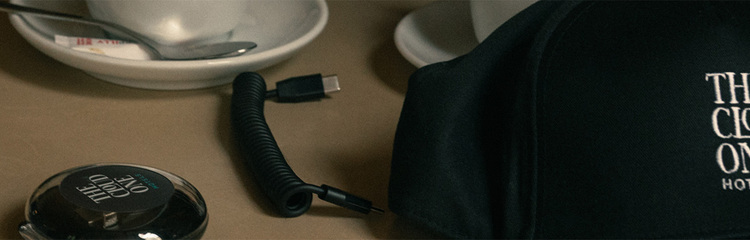 Eine schwarze Baseballkappe mit dem Schriftzug „THE CLOUD ONE HOTELS“ liegt auf einem Tisch, umgeben von persönlichen Gegenständen wie einem Smartphone, einem Kompass und Kopfhörern, die eine entspannte Reise- oder Urlaubsatmosphäre vermitteln.