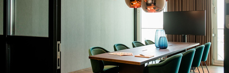 Moderner Konferenzraum mit großem Holztisch, acht grünen Stühlen und natürlichem Tageslicht durch große Fenster, ideal für Geschäftstreffen und Besprechungen.